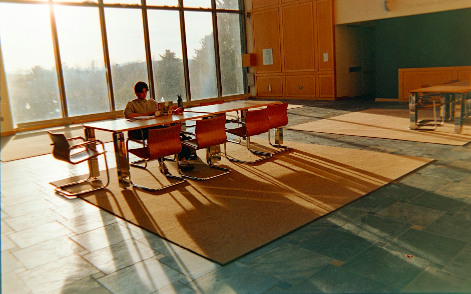 Una persona trabaja sola en una mesa larga, en un salón amplio con luz cálida de la tarde.